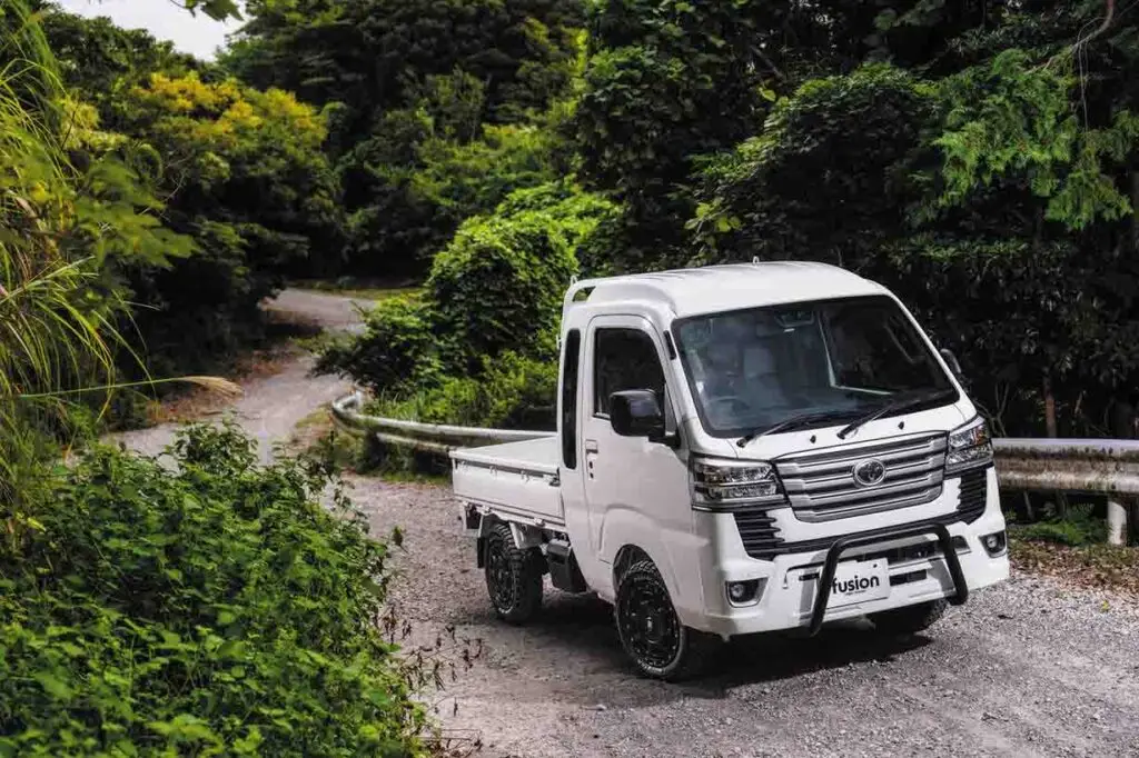 White customized Toyota Pixis Truck parked on a gravel path beside a guardrail in a forested winding road setting