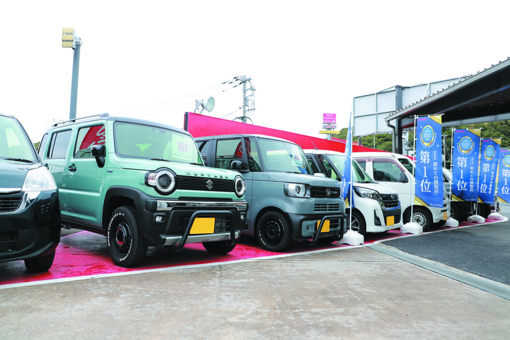 Real scene from a Japanese used vehicle dealership in the countryside showing kei cars including a Suzuki Hustler and Honda N-VAN lined up on a red display lot — the type of local shop where kei trucks are also traded
