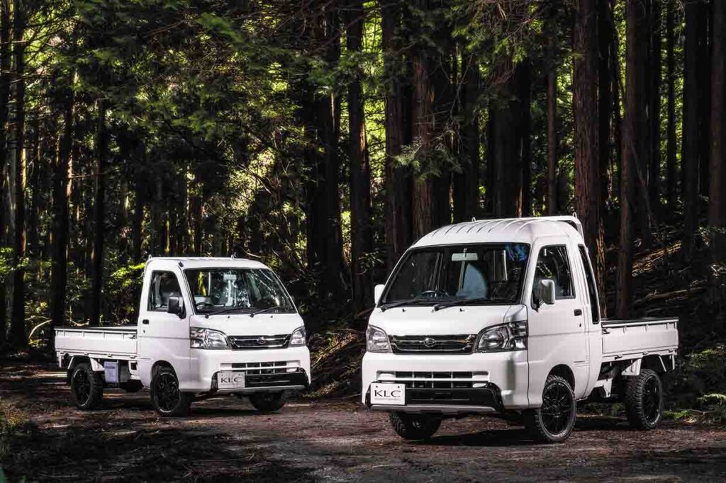Two white Daihatsu Hijet and Suzuki Carry kei trucks with KLC OFF BEAT front underguards parked side by side in a forest setting