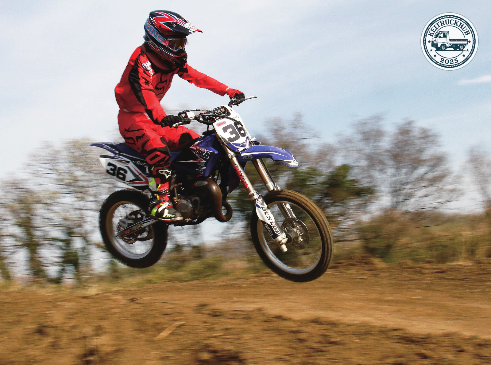 A helmeted rider in red gear rides the blue number 36 motocross bike over a rise on the dirt track.