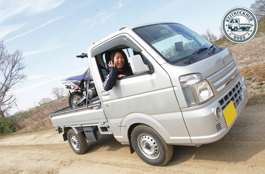 Shimizu waves from the open window of a silver kei truck with a blue motocross bike in its bed.